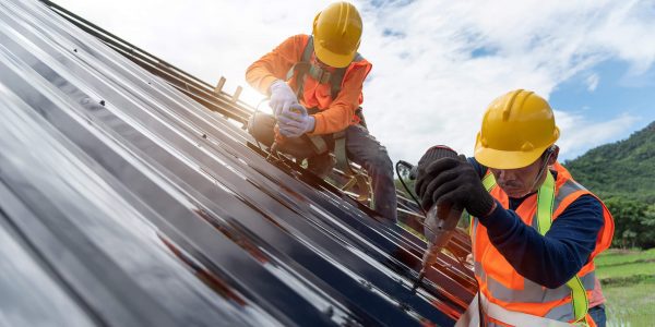 Two construction workers wearing orange safety vests and yellow helmets are busy with metal roofing tasks. One is drilling while the other secures materials, showcasing their expertise as roofing contractors. Above, the sky is partly cloudy, with verdant hills providing a picturesque backdrop.
