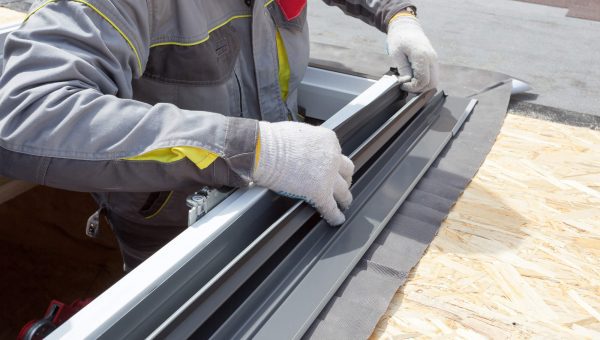 A roofing contractor in gray protective clothing and gloves expertly handles tools while installing a skylight window frame on a wooden roof surface. The scene captures the intricacy involved in skylight replacement, focusing on the skilled hands and precision of the process.