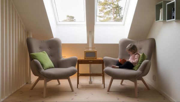 A child sits on a cozy armchair reading a book in a bright attic room with two skylights, recently attended to by a skilled roofing contractor. A small wooden table holds a radio between two armchairs, each with a green cushion. Neutral tones and a tree view create a peaceful atmosphere.