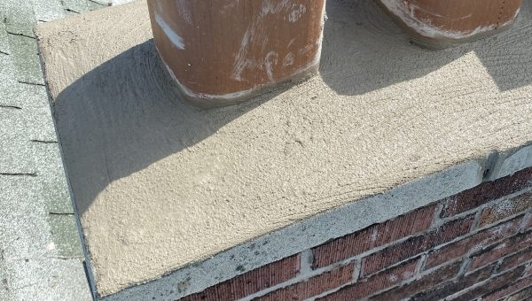 A brick chimney with two clay flue liners is under construction by a roofing contractor. The top is freshly cemented. Nearby, a bucket with tools and a tarp with roofing materials are visible. Roof shingles surround the chimney, highlighting the repair work in progress.