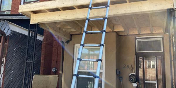 A person stands on a wooden balcony under construction on the second floor of a house, where a roofing company is busy with skylight repair. A long ladder leans against the balcony, reaching to the ground. The house features brick and beige siding near the entrance.