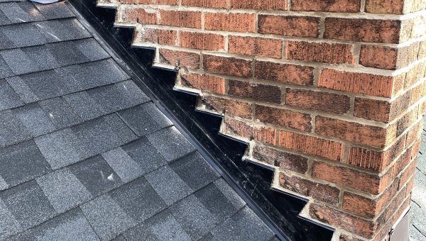 Close-up of a roof with dark shingles meeting a brick chimney. Black metal step flashing is visible along the side, ensuring weatherproof protection where the roof and chimney intersect—essential for preventing leaks during roof repair.