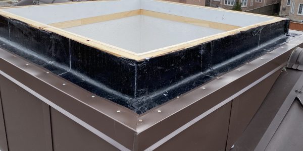 A rooftop skylight replacement is underway, featuring a wooden frame and black waterproof barrier, set against a backdrop of residential houses and a cloudy sky at sunset.