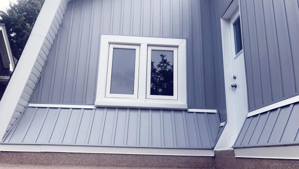 Modern building exterior with light grey metal siding and a sloped design, showcasing the expertise of a skilled roofing contractor. It features a white-framed window reflecting trees and a white door with a metal handle. The paved surface in front complements the tree visible on the left.