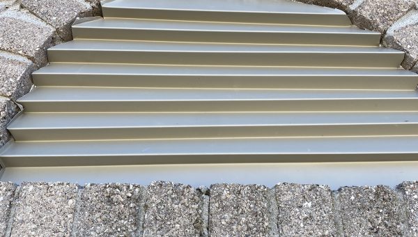 Close-up of a brick wall with an arched vent. The vent, crafted by a skilled roofing contractor, features horizontal metal slats and is framed by a stone arch made of bricks. The wall and arch are both textured with light-colored stones, showing expert workmanship.