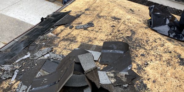 A rooftop with damaged and torn shingles scattered across a wooden surface. A residential street is visible with parked cars, a bicycle, and a recycling bin in the background. Consider calling a roofing company for skylight repair and metal roofing to enhance durability.