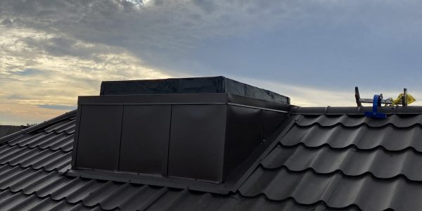 A rooftop view showcases dark gray tiles and a rectangular vent. The sky is cloudy, with sunlight peeking through, as tools resting on the tiles hint at ongoing roof repair by a skilled roofing company.
