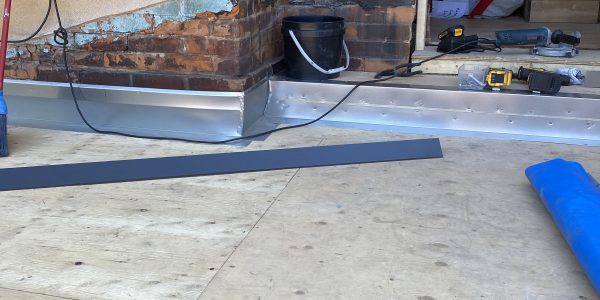 A partially renovated room with exposed brick and wood panels hints at ongoing roofing work. Tools and construction materials, including a power tool, screws, and a bucket, are scattered around. A blue mat and a long black strip are on the floor. A door opens to another room.