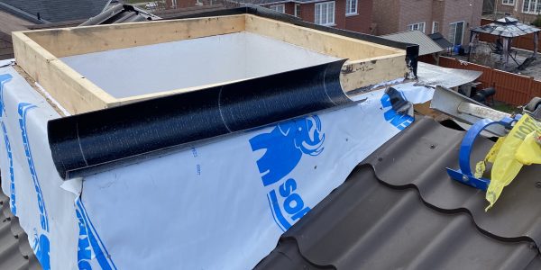 Rooftop with a partially constructed skylight. The frame is surrounded by waterproofing material with a blue logo. Nearby houses are visible under a cloudy sky. A roofing contractors tools and materials are scattered on the brown roof tiles, ready for skylight replacement.