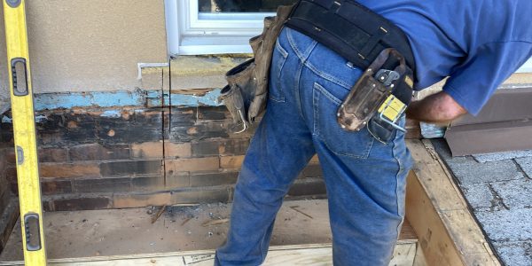 A construction worker in a blue shirt and jeans is measuring wood on a building site for a roofing contractor. A level tool is propped against the wall next to him. In the background, a window reflects two people and nearby buildings.