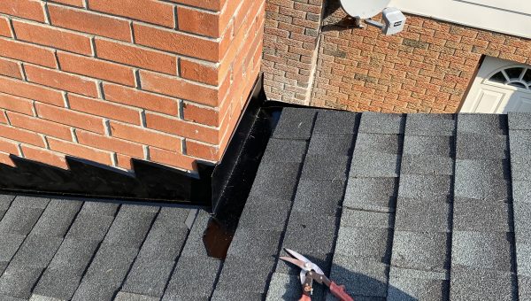Close-up of a rooftop showing a brick chimney with black flashing at the base, expertly installed by a roofing contractor. A pair of red-handled pliers lies on the gray shingles. In the background, theres a satellite dish attached to a brick wall, marked with Bell.