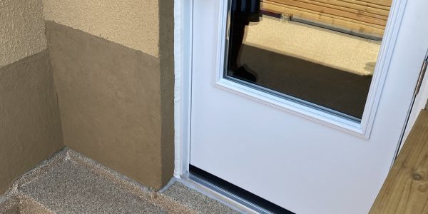 Exterior view of a white door with a reflective glass panel, set in a tan wall. The door opens to a small patio with wooden steps, perfect for contemplating roof repairs or consulting with your trusted roofing contractor. Reflections reveal nearby buildings and part of a person.