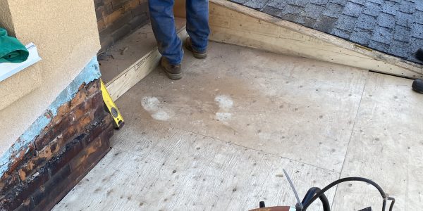 A construction worker wearing blue jeans and a blue shirt is focused on a skylight repair on a rooftop near a window. A saw is placed on a plywood surface. The area is under renovation, with tools and materials scattered around. The brick building features brown siding.