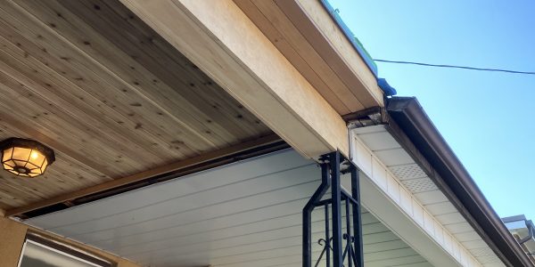 View from below of a house porch featuring distinct roofing styles—wood-paneled on the left and sleek white metal on the right, akin to designs a top roofing company might create. Black metal supports frame a wall with a window and light fixture, set against a clear blue sky above.