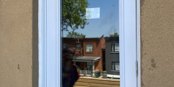 A glass door with a white frame reflects a person standing outside, beneath the flawless sky—a perfect day for roofing projects. Behind them, a wooden fence and townhouses complete the scene, with tree branches reaching towards the clear blue above.
