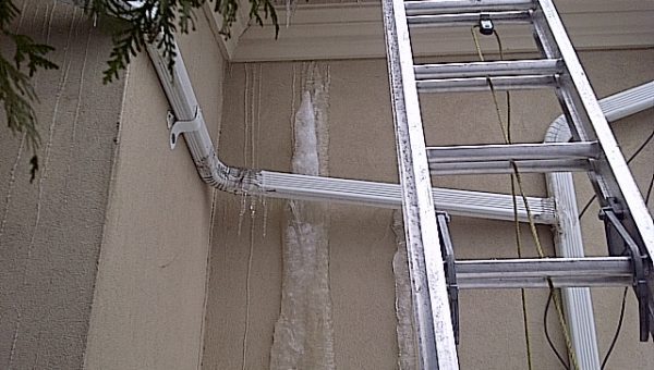 A metallic ladder leans against a building wall, indicating roofing activity. The wall features a gutter system with icicles hanging down, a sign of cold weather. A tree branch slightly extends into the top left corner of the image, capturing the wintry ambiance.