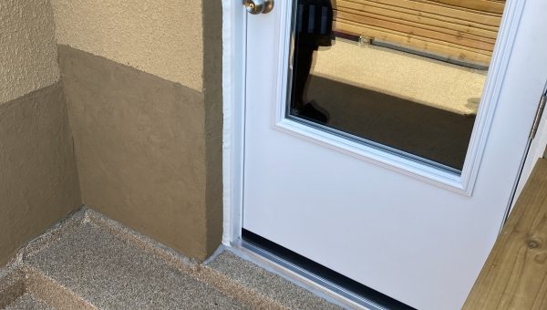 A white door with a glass window, reflecting nearby roofing company buildings, leads to a small outdoor landing. The landing has steps down to a wooden fence, offering a view of brick buildings in the background.