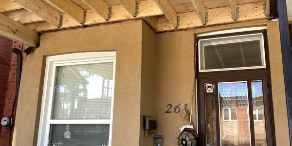 A house entrance with a wooden awning under construction showcases the meticulous work of a skilled roofing company. Theres a window and tall door numbered 261. A blue snow shovel leans against the wall near a bench, amid construction materials and tools.