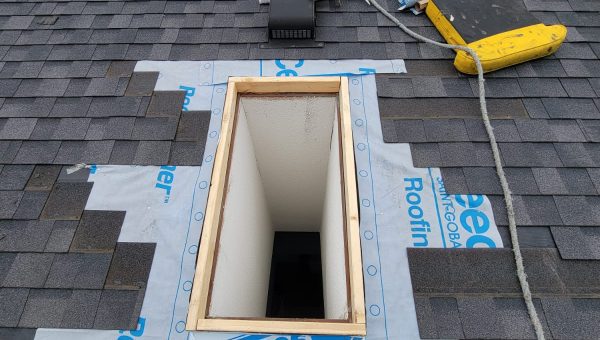 A roof prepared for skylight replacement features a rectangular hole surrounded by shingles and waterproofing tape. Tools, including a drill and caulk gun, signal ongoing roofing work. In the background, trees sway under a cloudy sky.