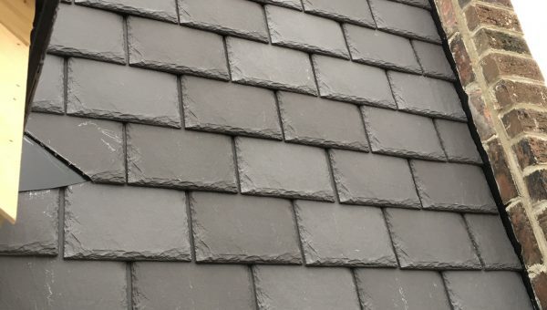 A close-up of a buildings exterior wall with dark gray slate tiles, reminiscent of precise roof repair work, arranged in a neat, overlapping pattern. The wall is adjacent to a section of exposed brickwork on the right.