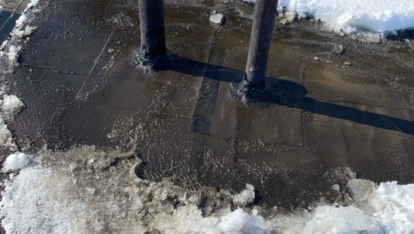A snow-covered roof with two vertical metal pipes protruding hints at the recent skylight repair. The surrounding snow is partially melted, revealing the dark surface beneath, typical of a professional roofing companys precise work. Shadows of the pipes are visible on the roof.