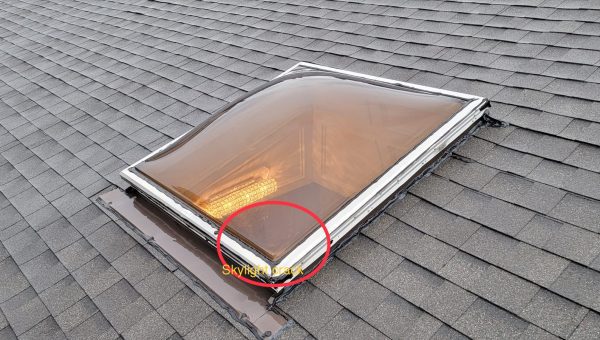 A curved skylight on a gray shingled roof displays condensation or fogging inside. A red circle and the word SKYLIGHT are overlaid near the corner of the skylight, highlighting the issue—a job for a skilled roofing contractor.