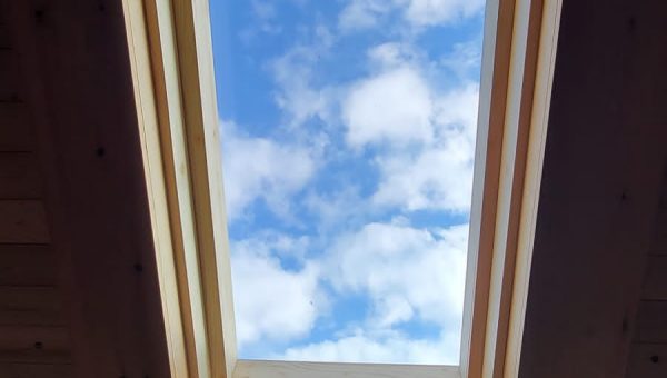 A rectangular skylight in a wooden ceiling frame reveals a view of a bright blue sky with scattered fluffy white clouds, showcasing the expert craftsmanship of our roofing company.