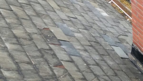 A close-up view of a slate roof with some damaged and missing tiles showcases the varied colors and textures. This weathered roof, captured by a roofing contractor, includes a partially visible brick chimney on the right edge of the image.