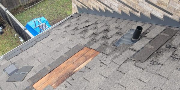 A person stands on a shingled roof with visible patches and a chimney vent, possibly assessing for skylight replacement. Nearby, a backyard below shows a blue container, grass, and a wooden fence. The adjacent buildings are made of beige brick.