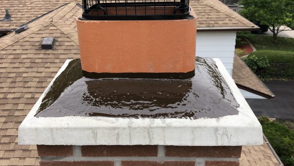 A brick chimney with a wet surface and a black metal chimney cap is seen atop the roofing. The roof of a house and surrounding greenery are visible in the background.