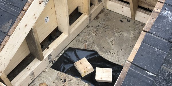 A rectangular hole in a roof, surrounded by wooden framing, likely for a skylight or chimney. The surrounding surface is covered in dark shingles, with two small wooden blocks resting inside the opening.