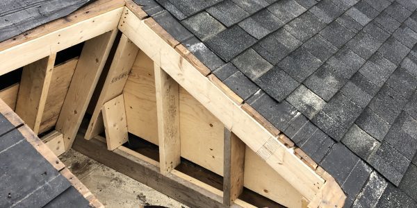 A roof with dark shingles partially removed, revealing wooden beams and an open section ready for repair or installation. A few yellow leaves are scattered on the shingles. Trees and part of a house are visible in the background.