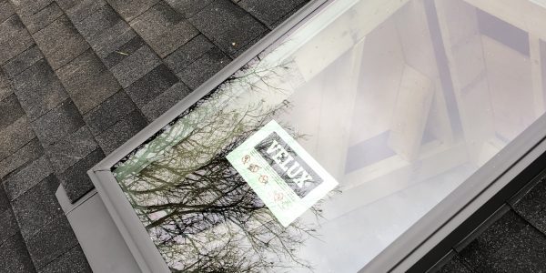 Skylight installed on a shingled roof, reflecting bare trees. A VELUX label is visible on the glass, courtesy of our skilled roofing contractor. Tools and roof sealant rest nearby.