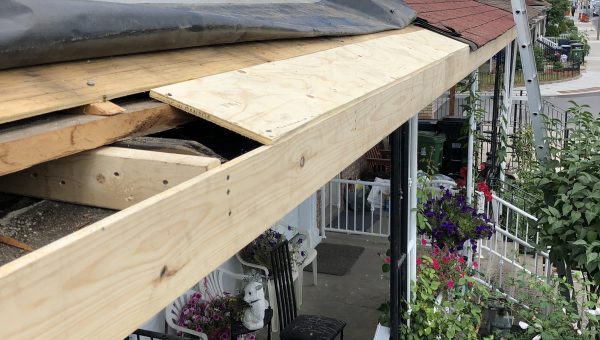 A partially repaired porch roof with exposed wooden beams and plywood signals the need for a roofing contractor. A black tarp covers part of the roof, while a ladder is propped against the building. Nearby, houses and plants accompany a trash bin and chairs on the porch.