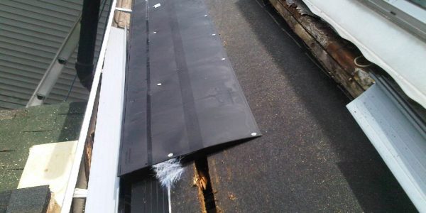 A roof with damaged roofing materials is partially covered by black sheets, showing signs of water damage and rot underneath. Nearby structures include a wall with windows and adjacent shingles. A roofing company could easily handle these repairs, ensuring durability and protection.