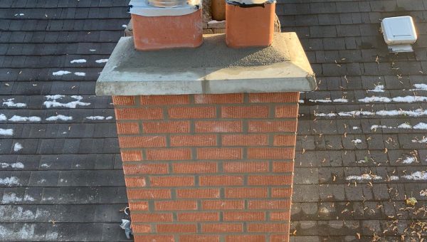 A brick chimney with two metal vents sits on a shingled rooftop, alongside a metal roofing section. Some patches of snow are visible on the roof. In the background, trees and other rooftops appear under a clear sky, hinting at possible skylight replacement needs.