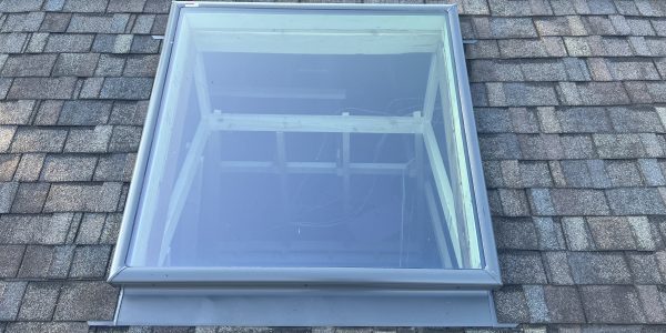 A skylight on a shingled rooftop, with a clear blue sky reflected in the glass, suggests recent roof repair. The surrounding shingles are various shades of gray and brown. Rooftops of other buildings and some greenery are visible in the background.