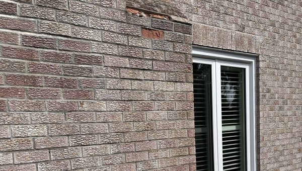 A beige brick wall with a rectangular patch of reddish bricks near the top left, and a white-framed double window with closed blinds on the right side. Sunlight casts shadows on the wall and window.