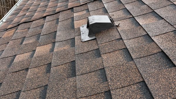 A close-up view of a sloped roof covered with brown asphalt shingles features two small metal vents. In the background, neighboring rooftops and a cloudy sky are visible, highlighting the craftsmanship of a top-notch roofing company.