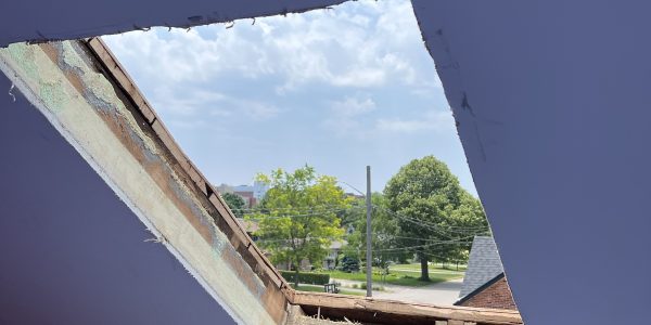 A triangular hole in a slanted ceiling reveals a view of a cloudy sky and treetops. The ceiling and walls, painted in shades of purple and pink, await attention from a roofing contractor to address the exposed insulation around the opening.