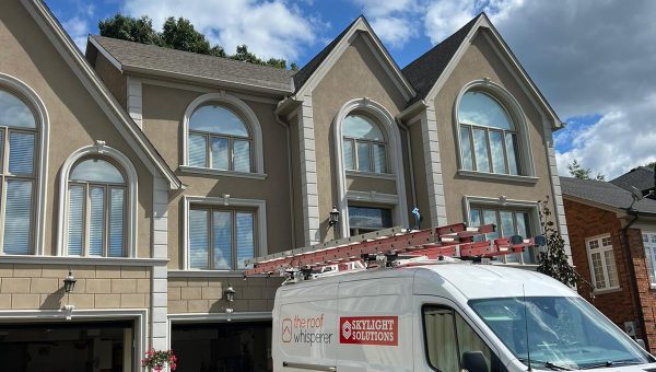 A white van with The Roof Whisperer and Skylight Solutions logos, a renowned roofing company, is parked in front of a large, beige house with multiple arched windows and a two-car garage. A red car is visible inside the garage. The sky is partly cloudy.