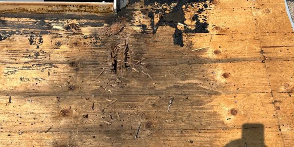 A section of the roof, expertly handled by a roofing company, reveals damaged plywood and removed nails and shingles. An open rectangular area suggests space for a skylight or entrance. Sunlight casts a shadow of a person holding a phone on the wood, capturing the roof repair process.