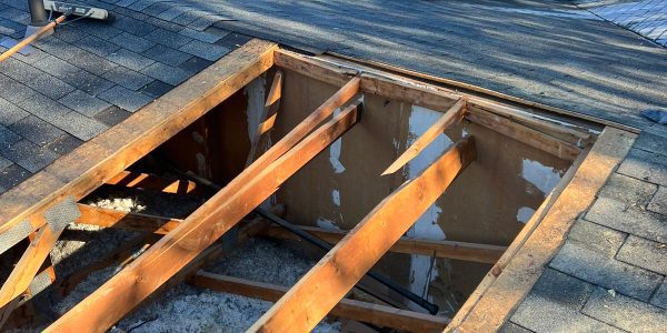 A house roof with an open section under repair, showing wooden beams and insulation, highlights the expertise of a roofing company. Nearby are tools like a shovel and hammer. Surrounding trees display autumn colors, with red and green foliage visible.