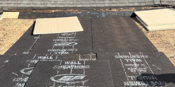 A rooftop under construction features dark roofing underlayment and two rectangular skylights in need of replacement. Nearby houses and a towering building form the backdrop against a cloud-dappled blue sky, as the roofing contractor meticulously transforms the skyline.