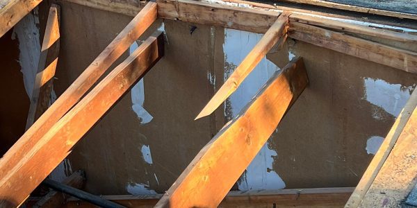 A view of an unfinished roof showing wooden beams and exposed insulation hints at a skylight replacement in progress. Part of the shingled roof is removed, revealing the interior structure and some tools. Sunlight casts shadows on the wooden beams and shingles.
