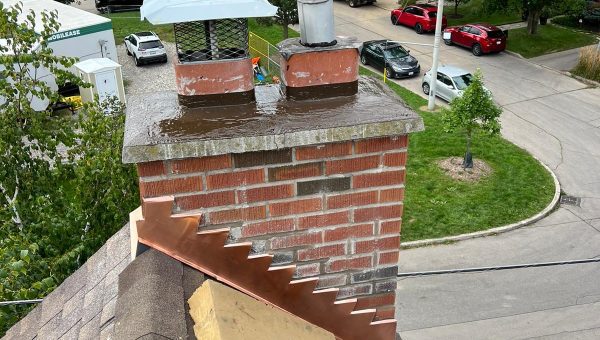 A rooftop scene showcases a brick chimney with a metallic cap and ventilation pipe. Copper flashing is expertly installed by a roofing contractor. Nearby, insulation material rests near the chimney. In the background, trees and parked vehicles line the residential street.