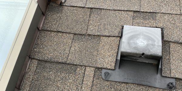 A section of a gray shingle roof, expertly handled by a roofing company, features a metal vent and adjacent skylight. The textured shingles and securely screwed vent cover enhance durability, while the skylights metal frame is ready for any needed skylight repair.