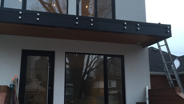 A modern two-story house with large windows and a flat roof is the perfect spot for skilled roof repair. The lower level features a black door, with containers and tools on the wooden steps leading up to it. A ladder rests against the right side, hinting at ongoing maintenance above.