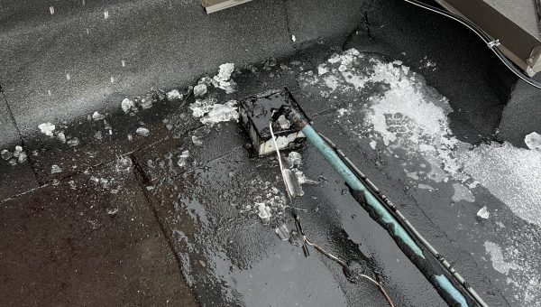 A rooftop with a puddle of water and patches of ice hints at the need for roof repair. Nearby, a small black box is connected to cables close to a blue pipe. A white service van from a local roofing company is parked on the street below, ready to address any issues.