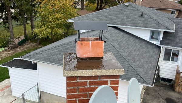 A rooftop view reveals a brick chimney topped with a wire-caged hood. Nearby, two satellite dishes are mounted, while skylight repair work is underway on the metal roofing. The backdrop features residential houses, vibrant autumn trees, and a cloudy sky.